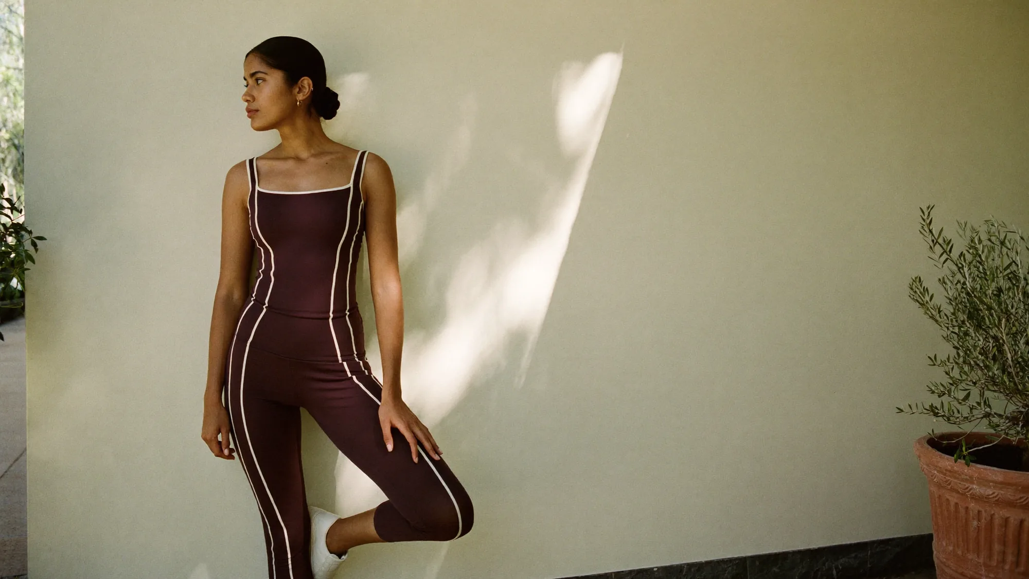 A woman in an aubergine tank and leggings with ivory piping, against a sunlit plaster wall.