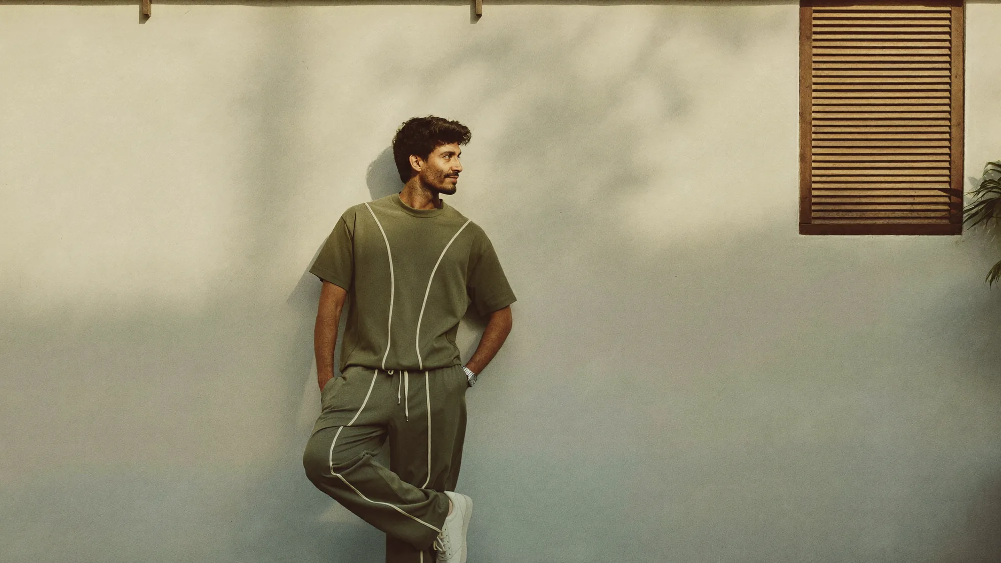 A man in an olive tee and wide-leg pants with ivory piping, against a sunlit plaster wall.